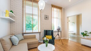 A modern living room with a sofa, a round coffee table with a vase of flowers, a dark armchair with a 'HAPPY' pillow, and large windows with blinds. - Bliss Apartments (Poznan)
