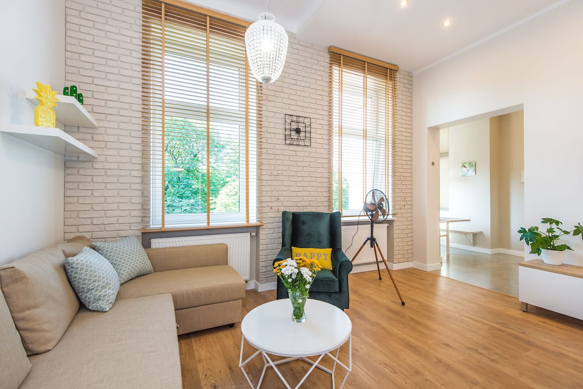 A modern living room with a sofa, a round coffee table with a vase of flowers, a dark armchair with a 'HAPPY' pillow, and large windows with blinds.