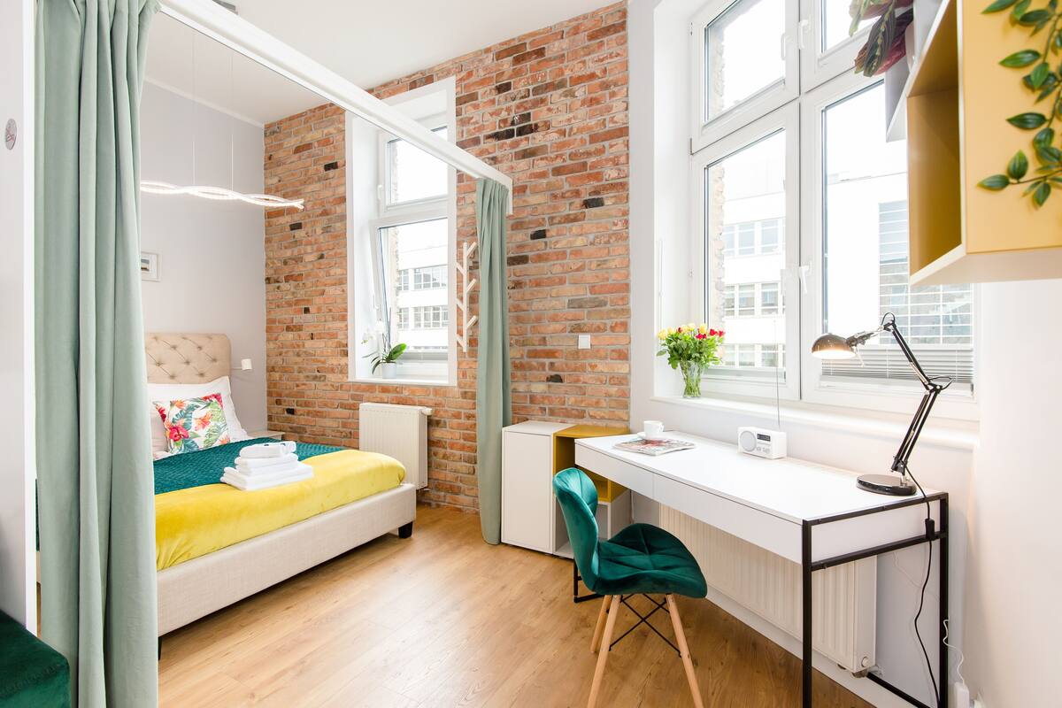 A modern bedroom with a brick wall, a bed with a yellow and green bedspread, a desk with a chair, and a window with a plant.