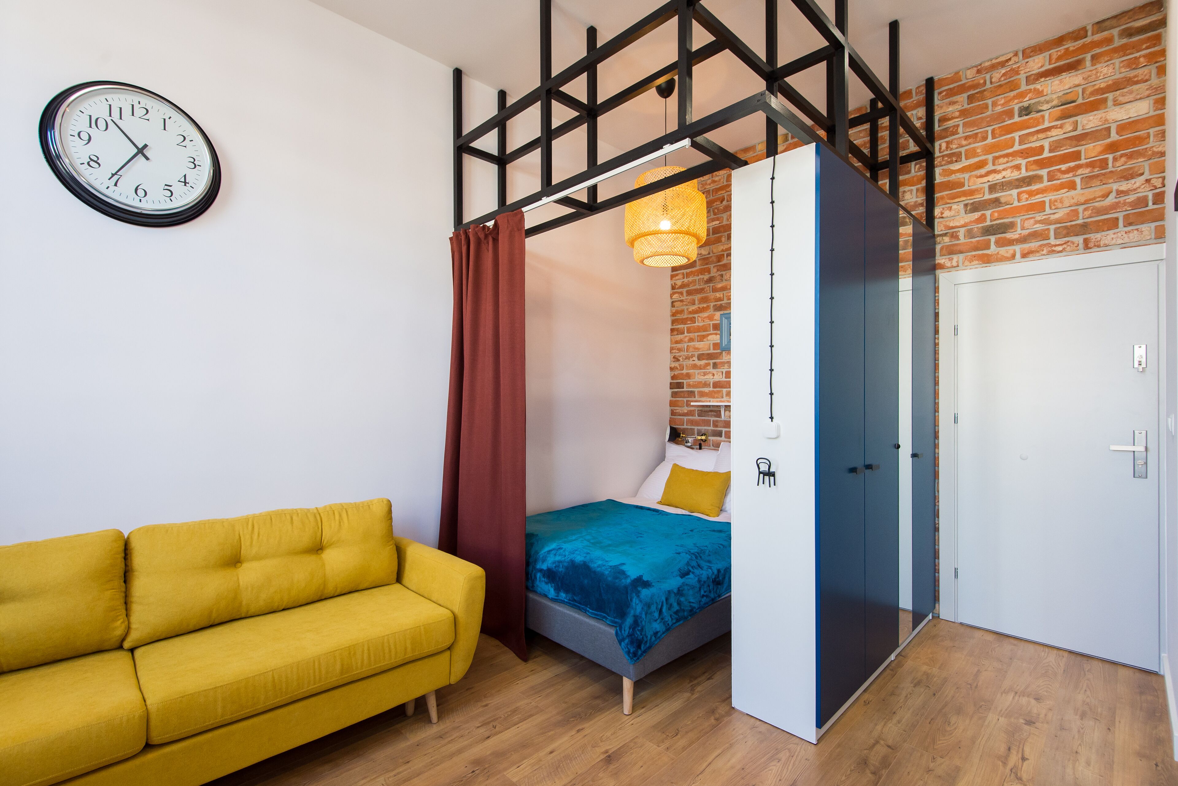 A room with a bed, a yellow sofa, a clock, and a brick wall.