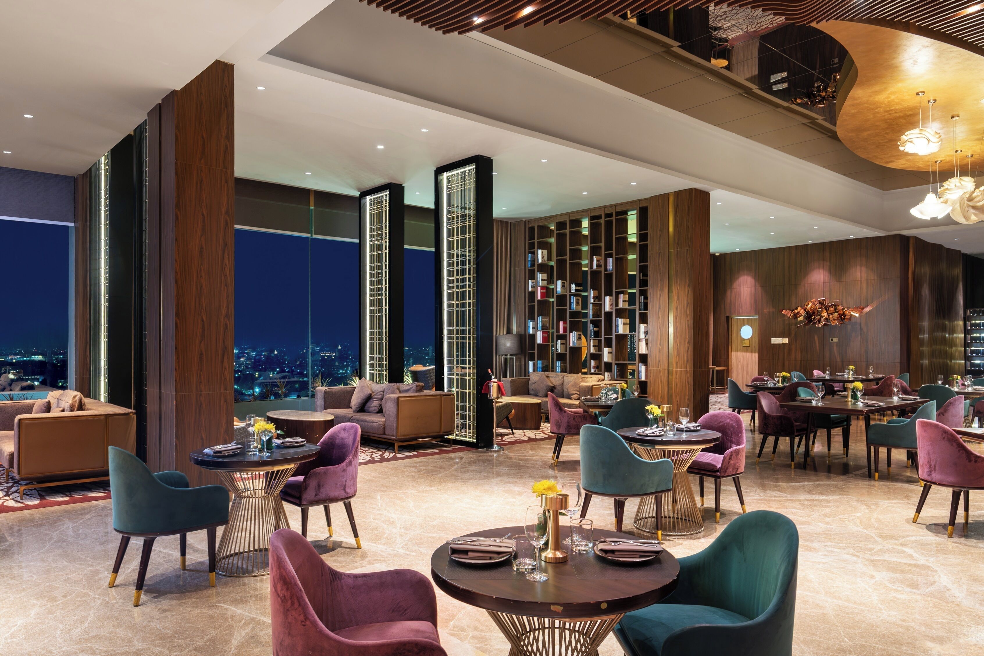 A spacious lobby with multiple seating areas, a large bookshelf, and a view of the city at night.