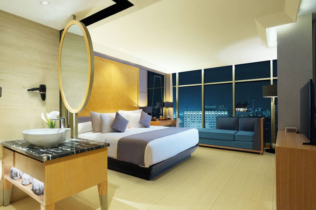 Ein modernes Hotelzimmer mit einem großen Bett, einem Badezimmer mit Marmorwaschtisch, einem großen Spiegel und Blick auf die Stadt durch die Fenster.