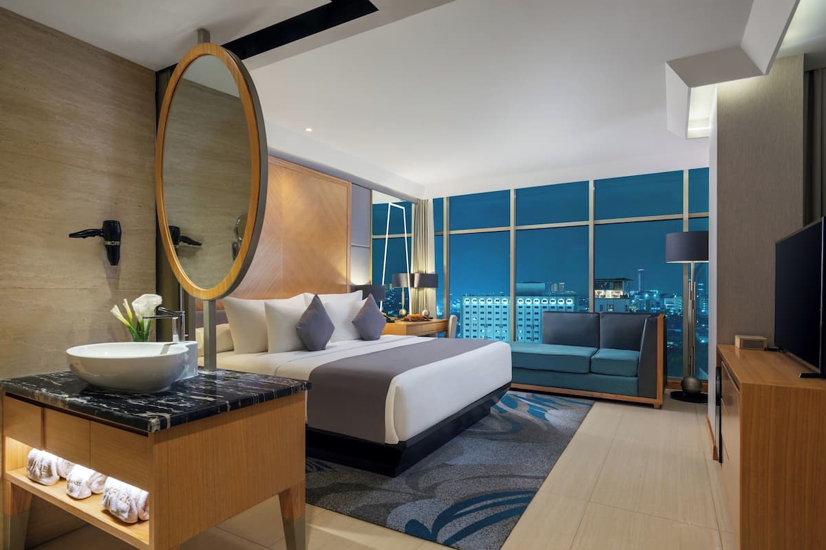Ein modernes Hotelzimmer mit einem großen Bett, einem runden Spiegel, einem Waschtisch mit einer Keramikwaschbecken und einem Flachbildfernseher, mit Blick auf die Stadt.