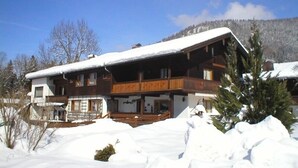 A two-story wooden chalet with a balcony, surrounded by snow-covered ground and trees. - Apartment Almrausch 75sqm, balcony, kitchen, 1 bedroom, 1 living room, WLAN (Ruhpolding)