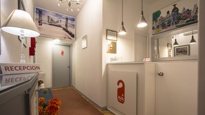 Zona de recepción de un hotel con un mostrador de recepción, un cartel que dice “RECEPCIÓN”, una lámpara de pared, un cuadro enmarcado y una puerta con un cartel que indica “2.0”.