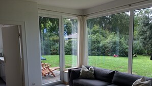 Ein modernes Wohnzimmer mit einem dunklen Sofa, einem Holztisch und Blick auf einen grünen Garten durch große Fenster.