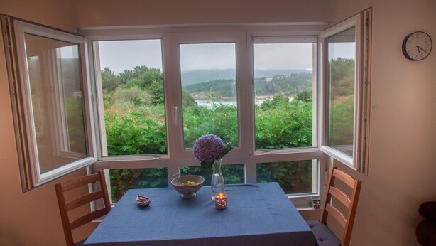 A dining area with a table set for two, a vase with flowers, and a view of greenery and hills through large windows. - Casa de Piedra Playa de Estorde con vistas al mar (Cee)