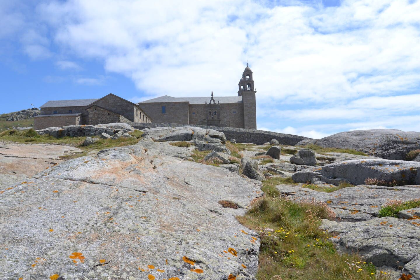 Un edificio de piedra con un campanario, rodeado de rocas grandes y vegetación escasa.