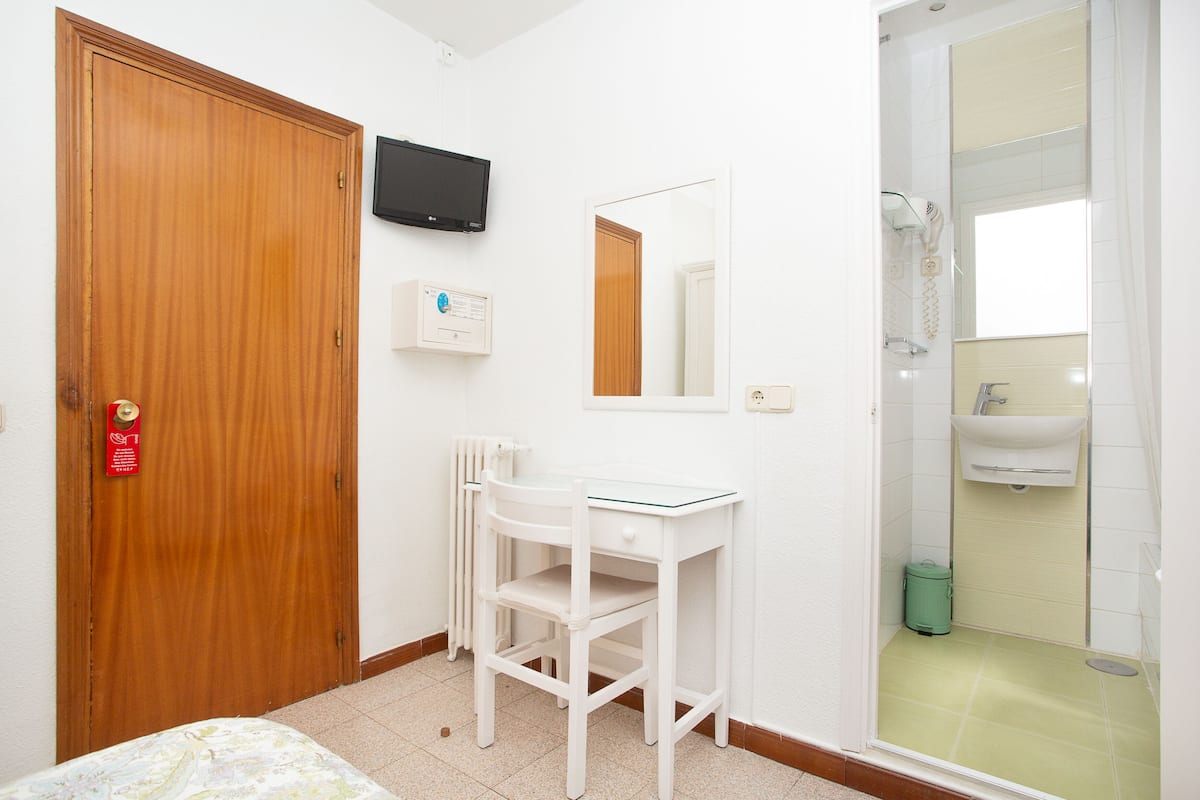 A hotel room with a wooden door, a white desk with drawers, a chair, a television mounted on the wall, a mirror, and a shower area with a sink.