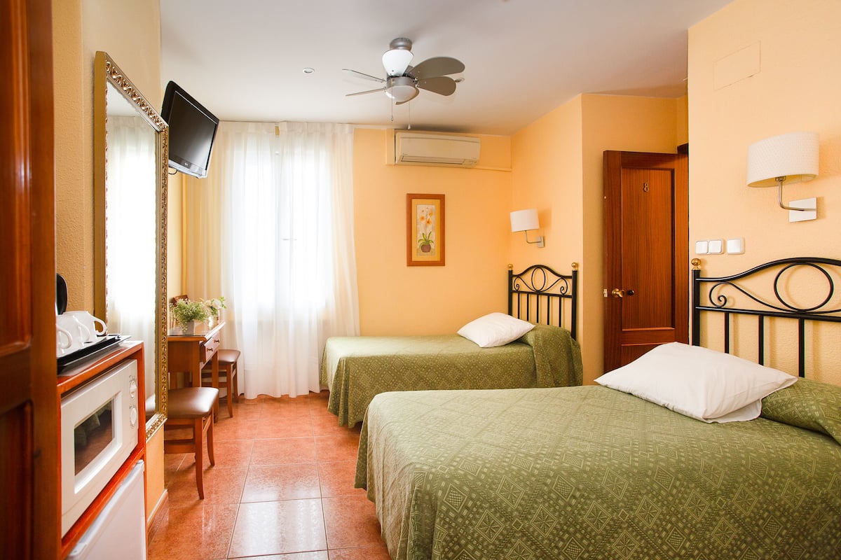 A hotel room with two beds, a TV mounted on the wall, a ceiling fan, an air conditioning unit, a wooden door, and a small desk with a chair.