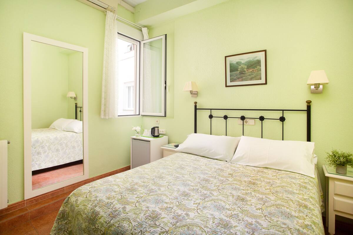A bedroom with a bed, a nightstand, a bedside table, a mirror, a window, and a wall-mounted picture.