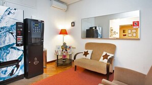 A compact living space with a sofa, a small table with a lamp, a refrigerator, and a mirror on the wall. - Hostal Los Alpes (Madrid)