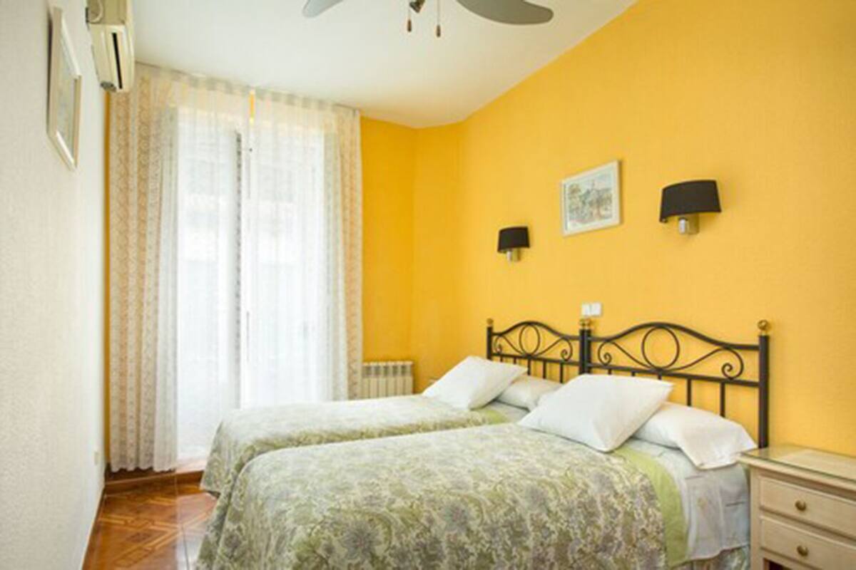 A room with two beds, a ceiling fan, a radiator, and a window with curtains.
