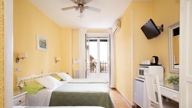 A hotel room with two beds, a desk, a chair, a television, and a balcony with a view. - Hostal Los Alpes (Madrid)