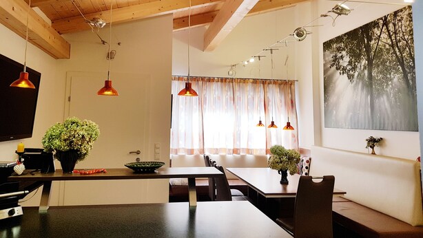 A modern kitchen with wooden beams, pendant lights, a dining area with a table and chairs, and a large window with curtains.