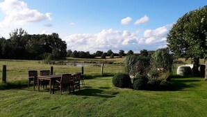 A grassy outdoor area with a table and chairs, a pond, and trees in the background.