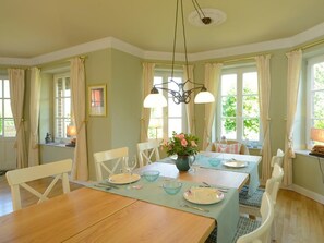 Ein Esszimmer mit Holztisch, gedeckt für vier Personen, weißen Stühlen, einer Pendelleuchte und großen Fenstern mit Vorhängen.