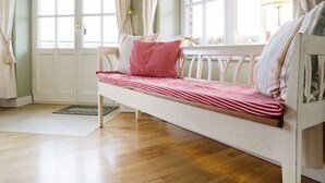 Quarto com piso de madeira, um banco branco com roupa de cama listrada e uma janela com cortinas.