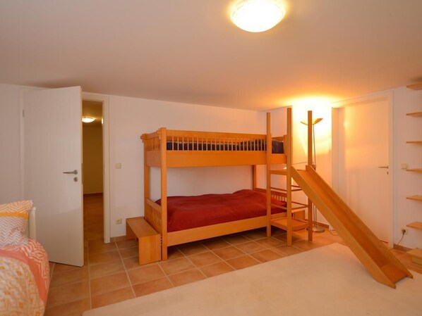 Una cama litera con escalera de madera y un pequeño tobogán, una puerta blanca y un suelo de baldosas.