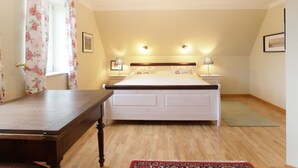 A bedroom with a wooden floor, a bed with white bedding, two bedside tables with lamps, a window with floral curtains, and a small rug.