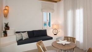 Una habitación con un sofá oscuro, una mesa redonda, sillas de mimbre, una lámpara y vistas a través de una ventana.