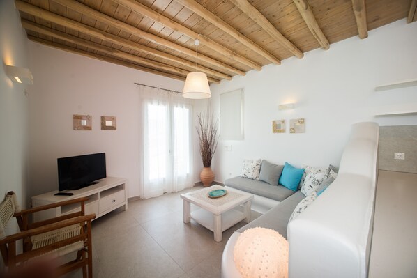 A living room with a wooden ceiling, a television on a white cabinet, a sofa with patterned cushions, and a coffee table.