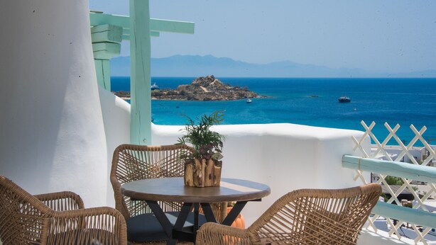 Un balcón con muebles de mimbre y una planta en maceta, con vista al mar, barcos y montañas a lo lejos.