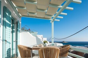 A balcony with a table and chairs, a white building with blue shutters, and a view of the sea.
