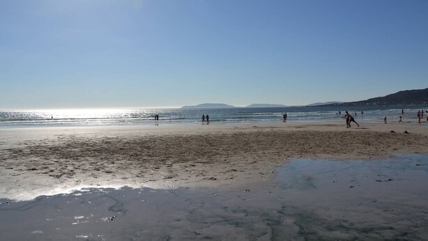 A beach with people walking and playing, a clear sky, and distant mountains.
