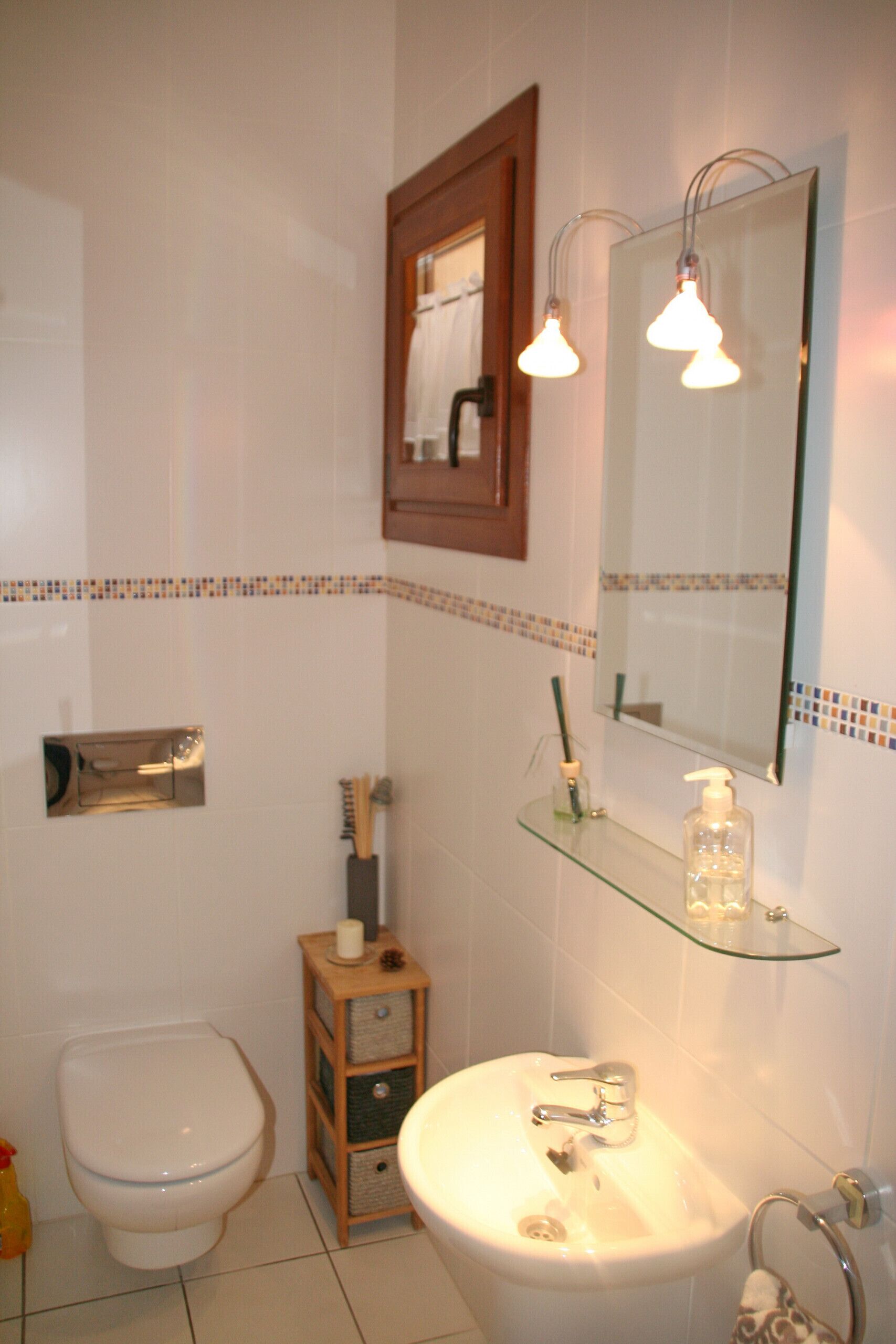 Une salle de bain comprenant des toilettes, un lavabo, un miroir et une étagère en verre où sont rangés les articles de toilette.