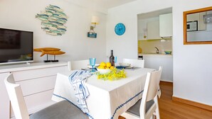 A dining area with a white table set for two, a fish wall art, a clock, and a kitchen in the background.