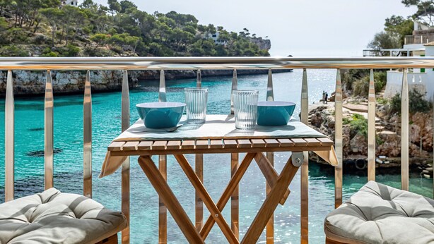 A wooden table with two blue bowls and two glasses, overlooking a turquoise sea with rocky cliffs and greenery.