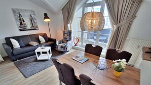 Een moderne woonkamer met een bank, salontafel en eettafel met stoelen. Er is een groot raam met gordijnen, een hanglamp en een schilderij aan de muur.