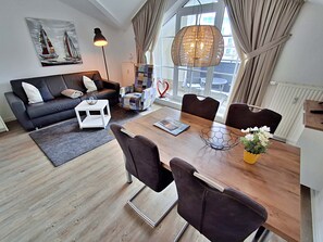 Un salon moderne comprenant un canapé, une table basse et une table à manger avec des chaises. On y trouve une grande fenêtre avec des rideaux, un luminaire suspendu et un tableau accroché au mur.