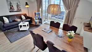 Een moderne woonkamer met een bank, salontafel en een eetgedeelte met stoelen. Er is een groot raam met gordijnen, een hanglamp en een boekenkast.