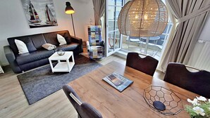 Een moderne woonkamer met een bank, salontafel, eettafel en stoelen. Er is een boekenkast, een lamp en een groot raam met gordijnen.