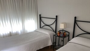 Deux lits simples avec du linge de lit blanc, une table de chevet avec une lampe et une fenêtre avec des rideaux légers.