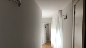 Un couloir étroit aux murs blancs et au plancher en bois mène à une porte ornée d’un rideau blanc.