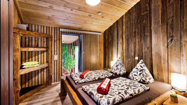 Interior de uma cabana de madeira com uma cama, almofadas, uma prateleira e uma pequena mesa com uma planta.
