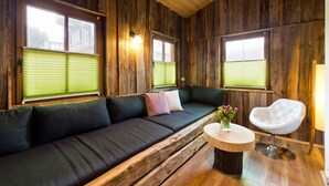 Um quarto com painéis de madeira, um sofá verde escuro, duas janelas com persianas, uma cadeira branca e uma pequena mesa redonda com um vaso de flores.