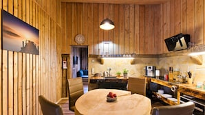Interior de madeira com uma mesa de jantar redonda, cadeiras, um relógio e uma área de cozinha com micro-ondas e cafeteira.