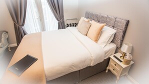 A neatly made bed with a laptop, a bedside table with a lamp, and a window with curtains.