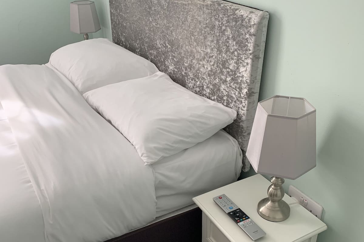 A neatly made bed with white linens, a grey textured headboard, a bedside lamp, and a remote control on a small white nightstand.