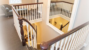 A staircase with a wooden handrail and white balusters, leading to a second-floor landing with a chandelier and a framed picture.
