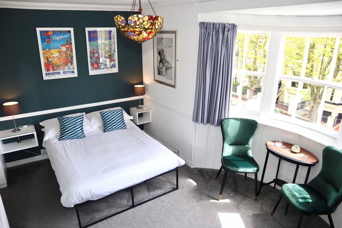 A bedroom with a bed, two chairs, a small table, a lamp, and a window with curtains.