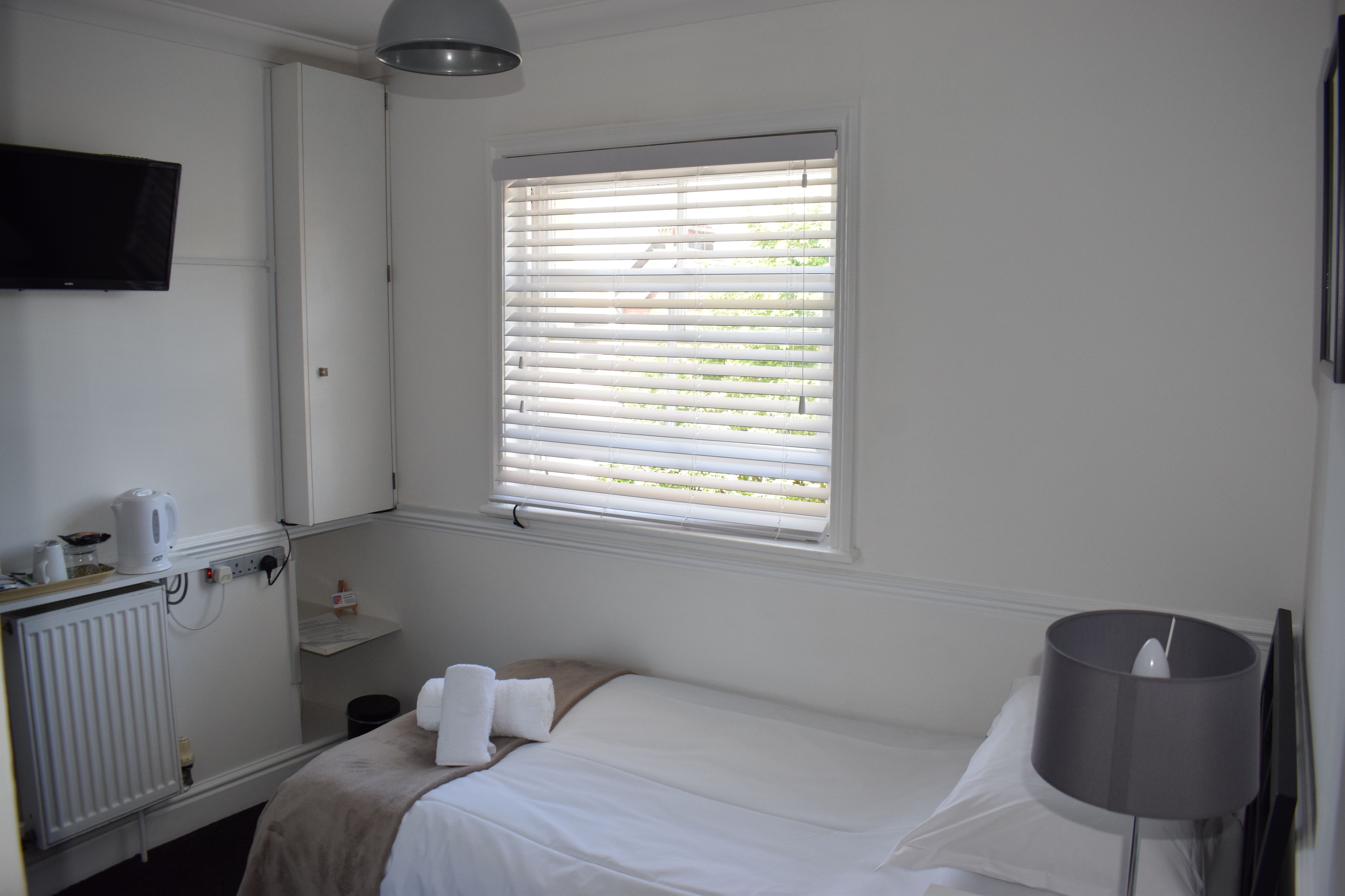 A hotel room with a bed, a bedside lamp, a window with blinds, a radiator, a TV mounted on the wall, and a small shelf with toiletries.