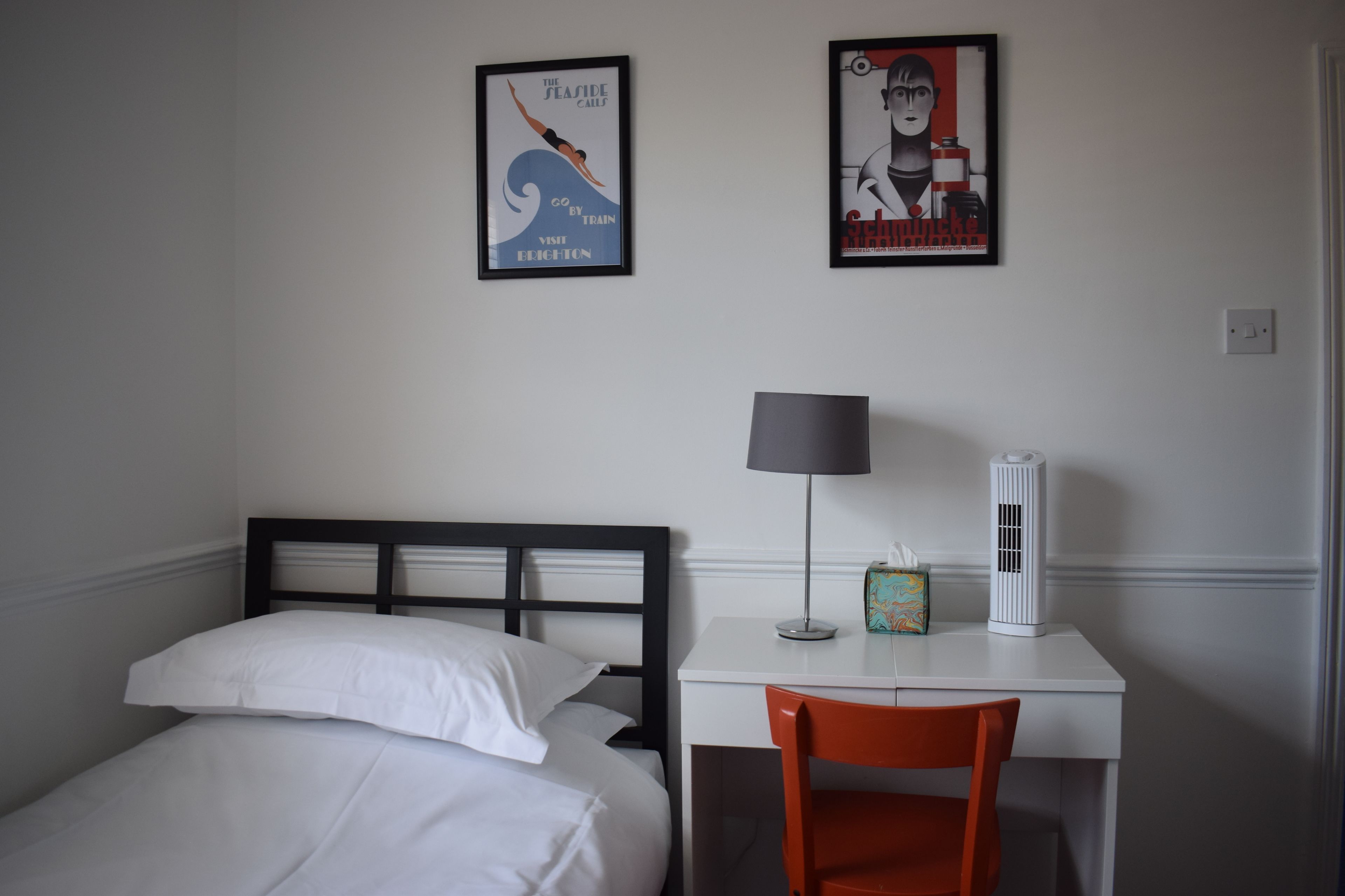 A hotel room with a bed, a desk, an orange chair, a lamp, and two framed posters on the wall.