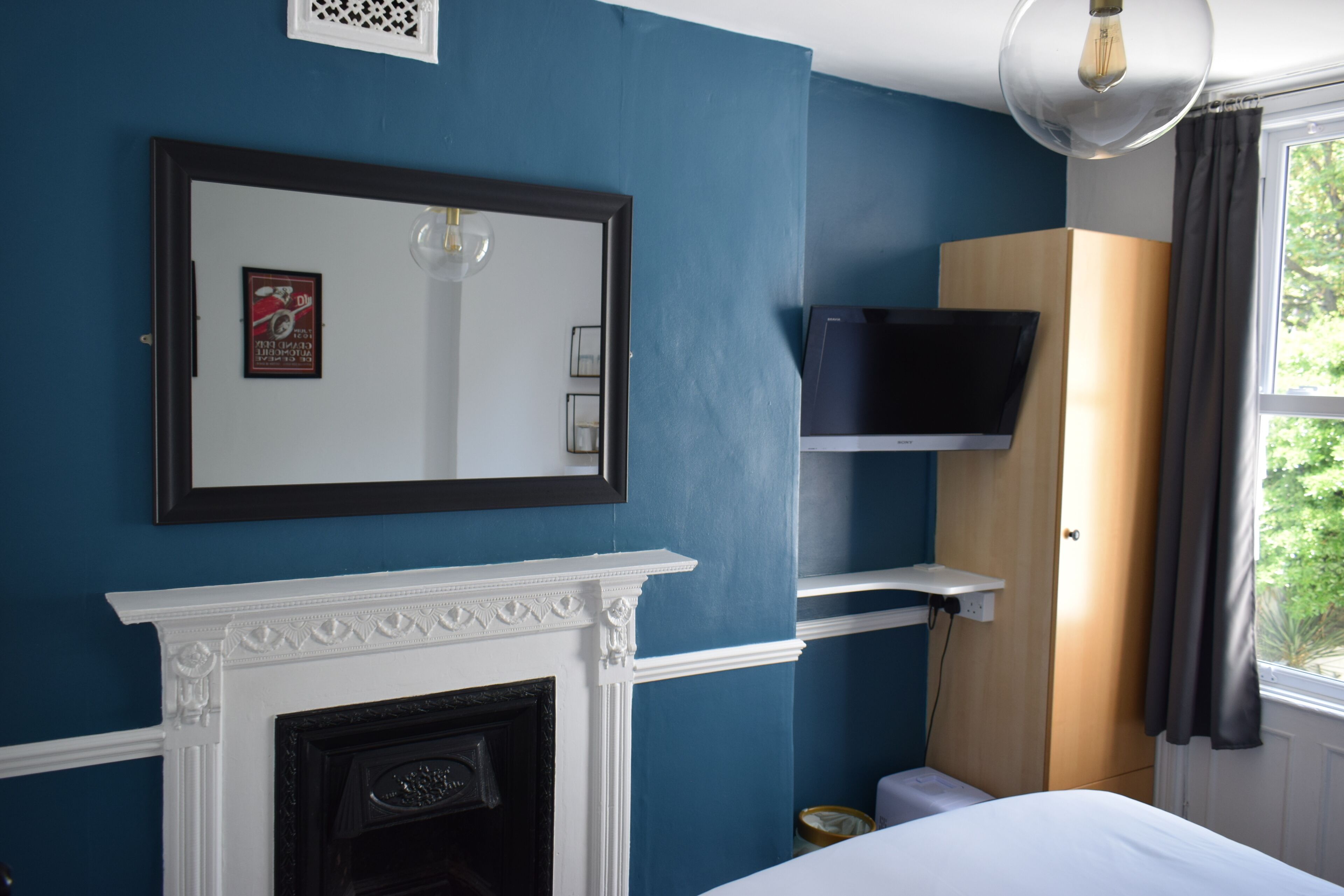 Un dormitorio con pared azul, chimenea blanca, televisor montado y ventana con cortinas.
