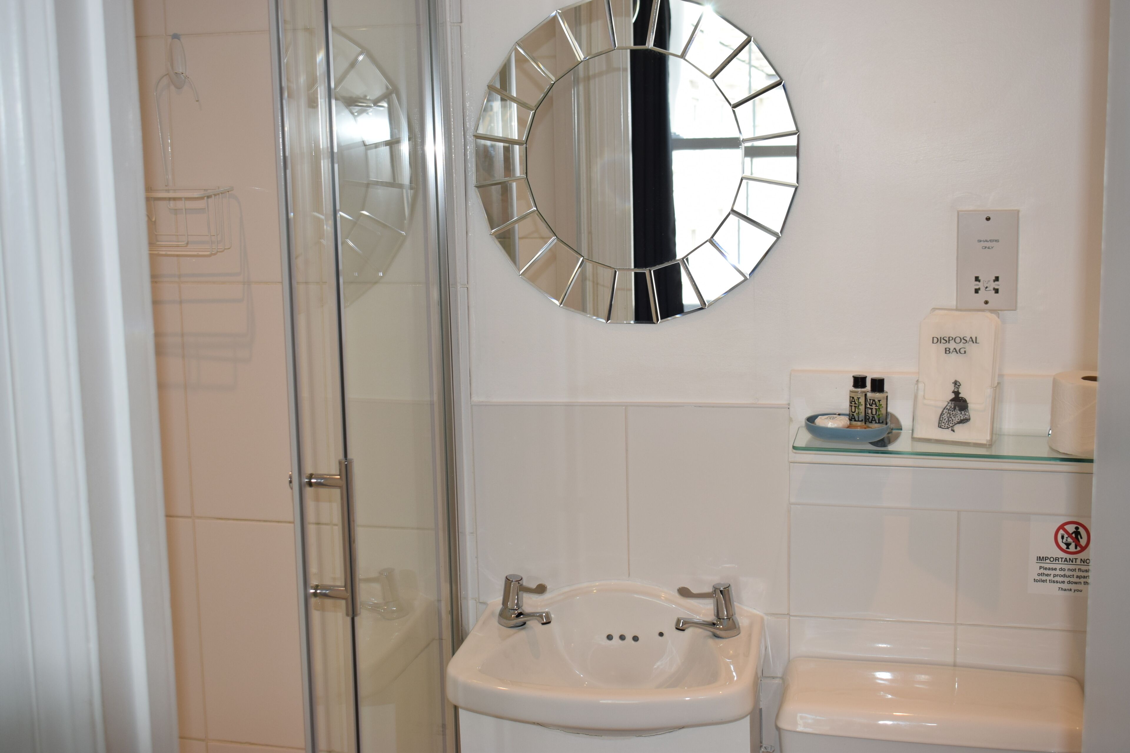 A bathroom with a glass shower enclosure, a sink with two faucets, a round mirror, and a toilet with a disposal bag.