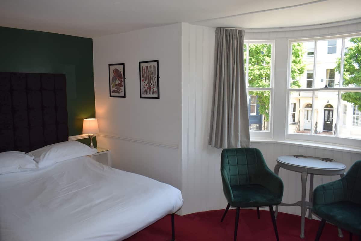 A hotel room with a bed, two chairs, a small table, and a window with a view of buildings and trees.